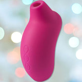 The LELO SONA Cerise - Sonic Clitoral Massager, crafted by LELO, is showcased with a waterproof design and side buttons against a softly blurred backdrop of pastel lights. The device's smooth, curved form enhances the experience through its innovative contactless Sonic Pulse technology.